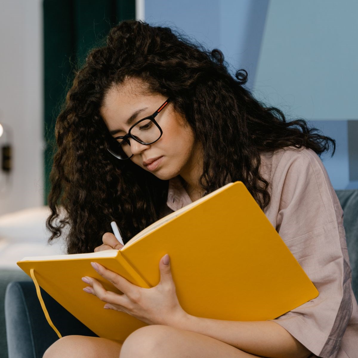 Young woman wearing glasses is sitting down, engrossed in writing her thoughts down in a journal