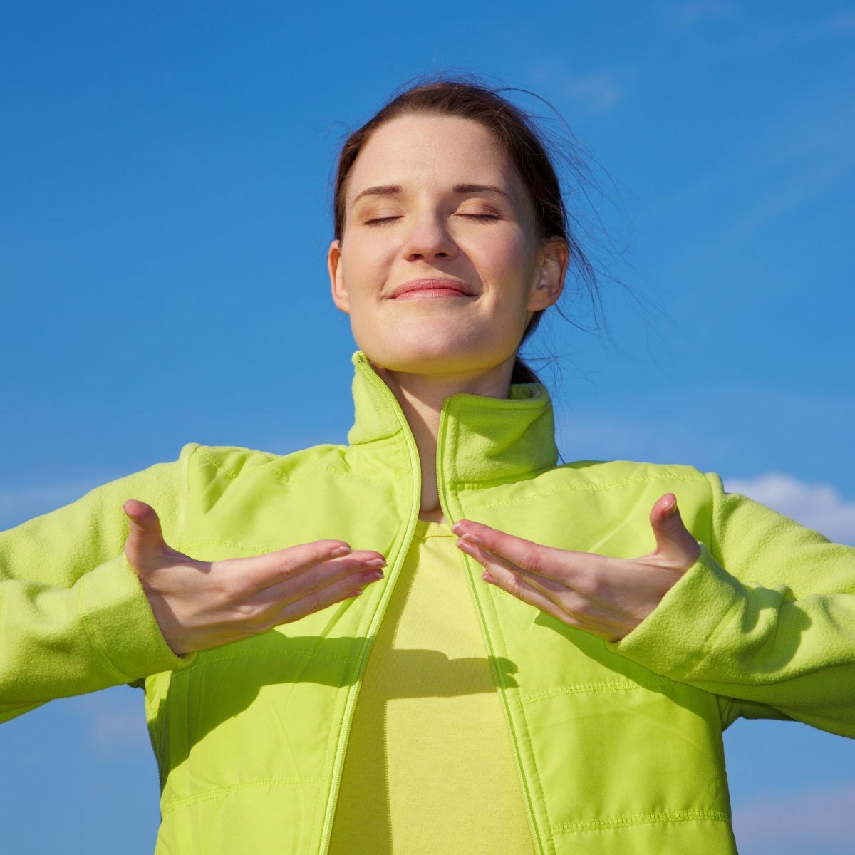 Young content-looking woman stands outside on a sunny day, closing her eyes while inhaling fresh air 