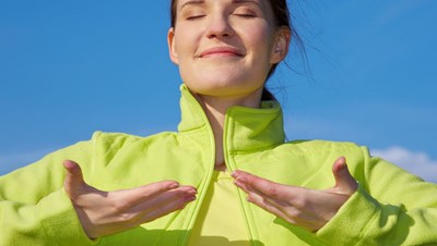Young content-looking woman stands outside on a sunny day, closing her eyes while inhaling fresh air 