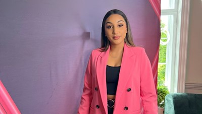Portrait photo of patient Nikita looking content. She is standing against a purple backdrop, and wearing pink jacket over blacktop and trousers